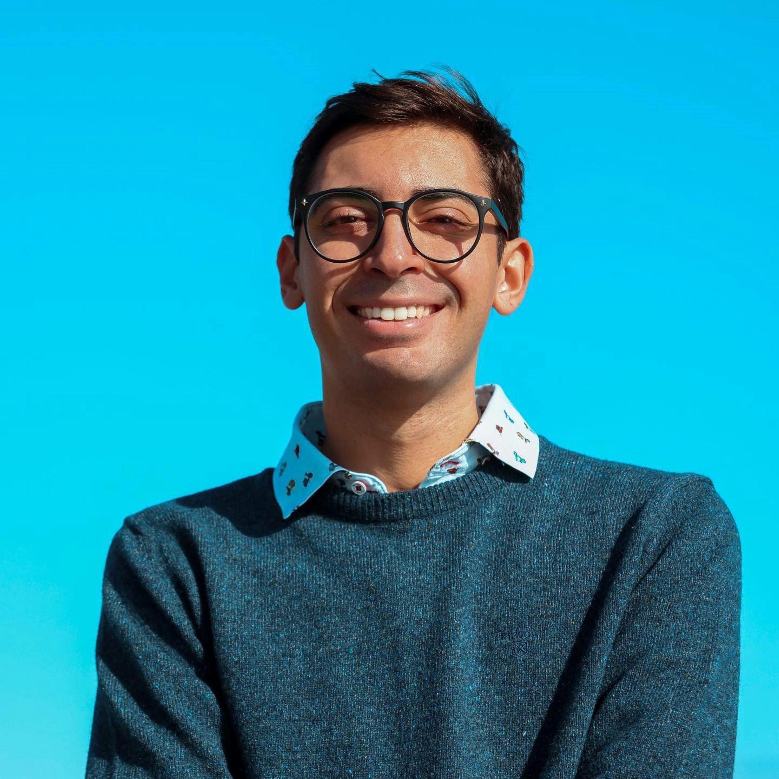 A portrait of a young man wearing stylish glasses and a blue sweater against a clear sky.